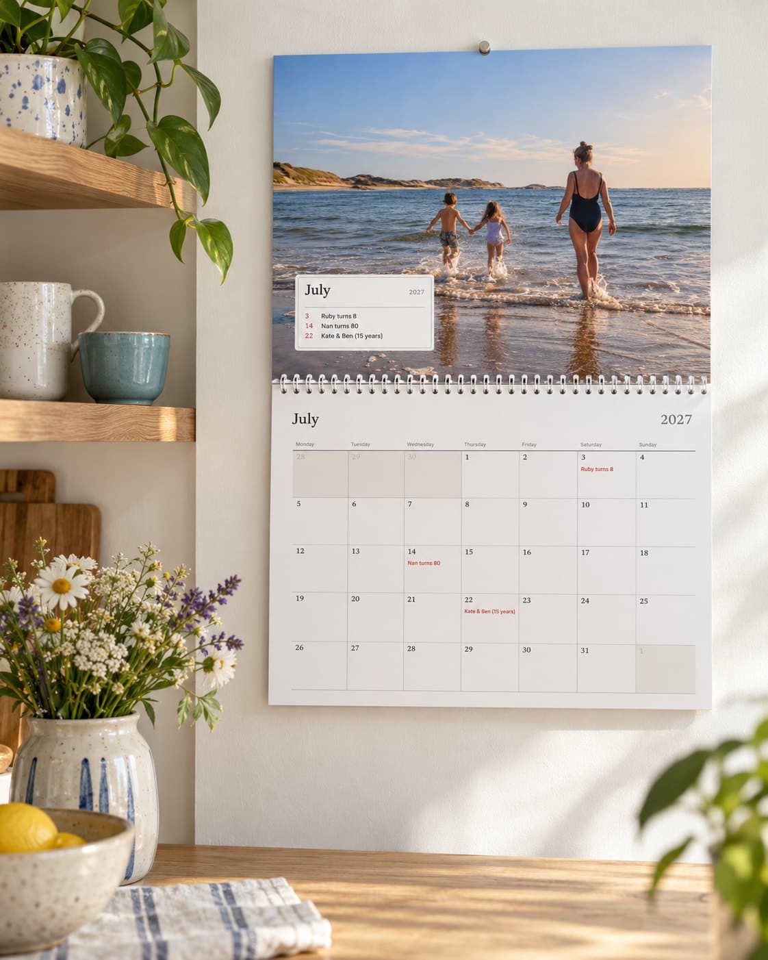 A photo calendar open at July in a bright kitchen: a family's beach photo above the month with their birthdays