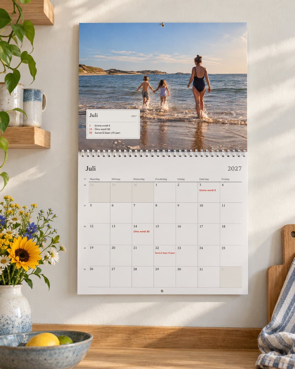 Een fotokalender open op juli in een lichte keuken: een strandfoto van het gezin boven de maand met hun verjaardagen