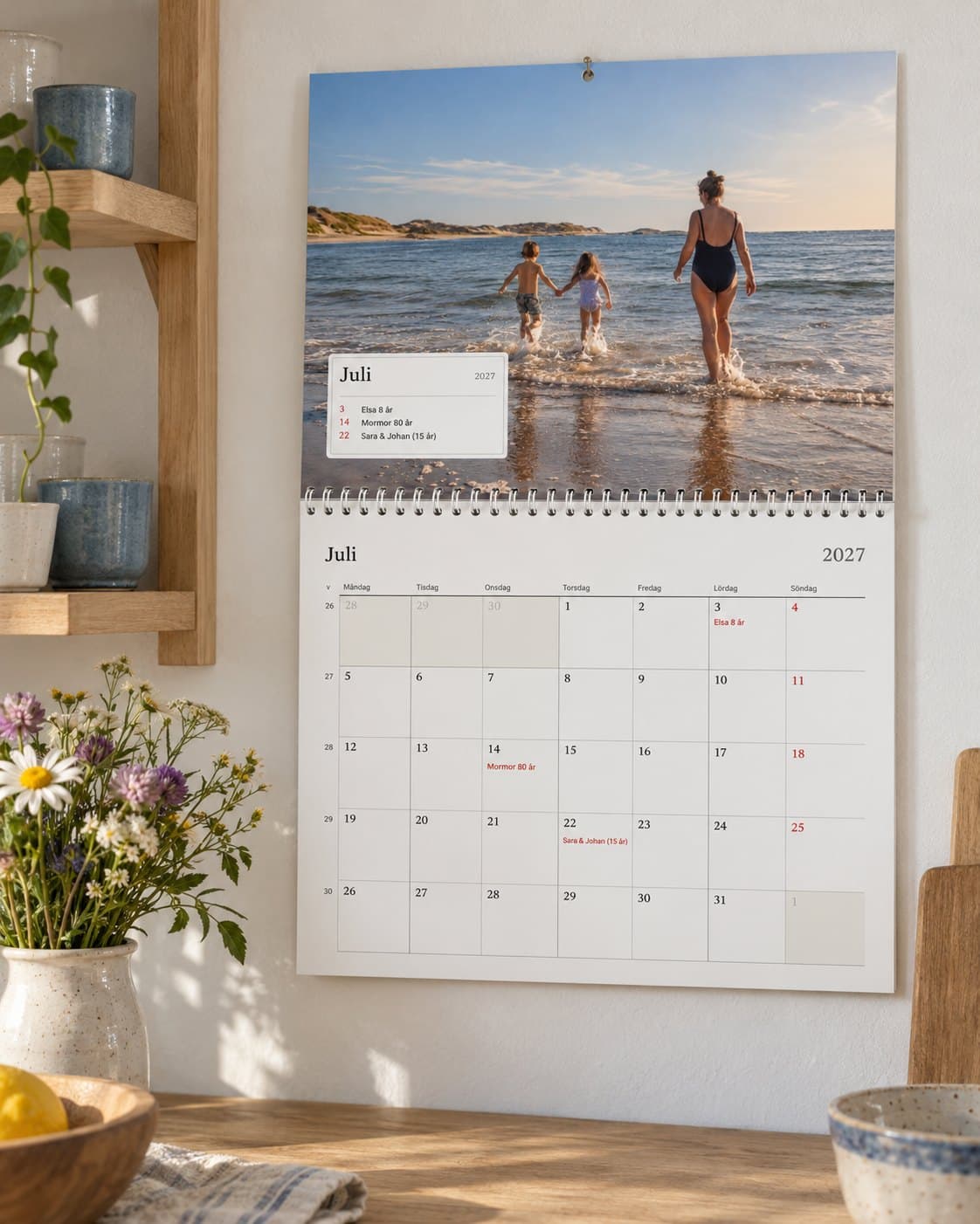 En fotokalender uppslagen på juli i ett ljust kök: familjens strandbild ovanför månaden med deras födelsedagar